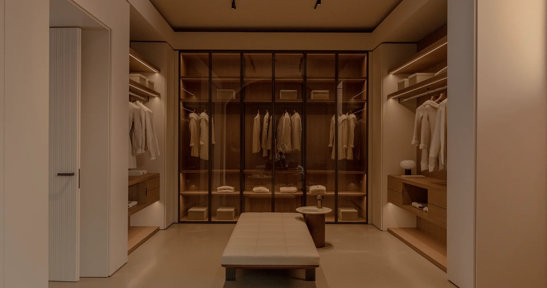 Bespoke dressing room with illuminated timber wardrobes, glazed doors and a central upholstered bench